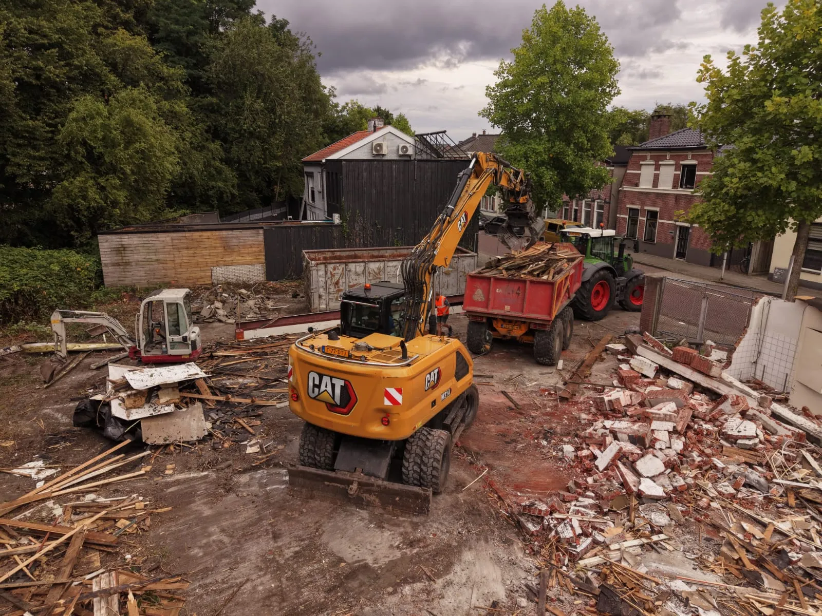 Een CAT graafmachine laadt bouwafval en hout in een rode aanhanger op een bouwwerf met puin en hout in een woonwijk met bomen en huizen.