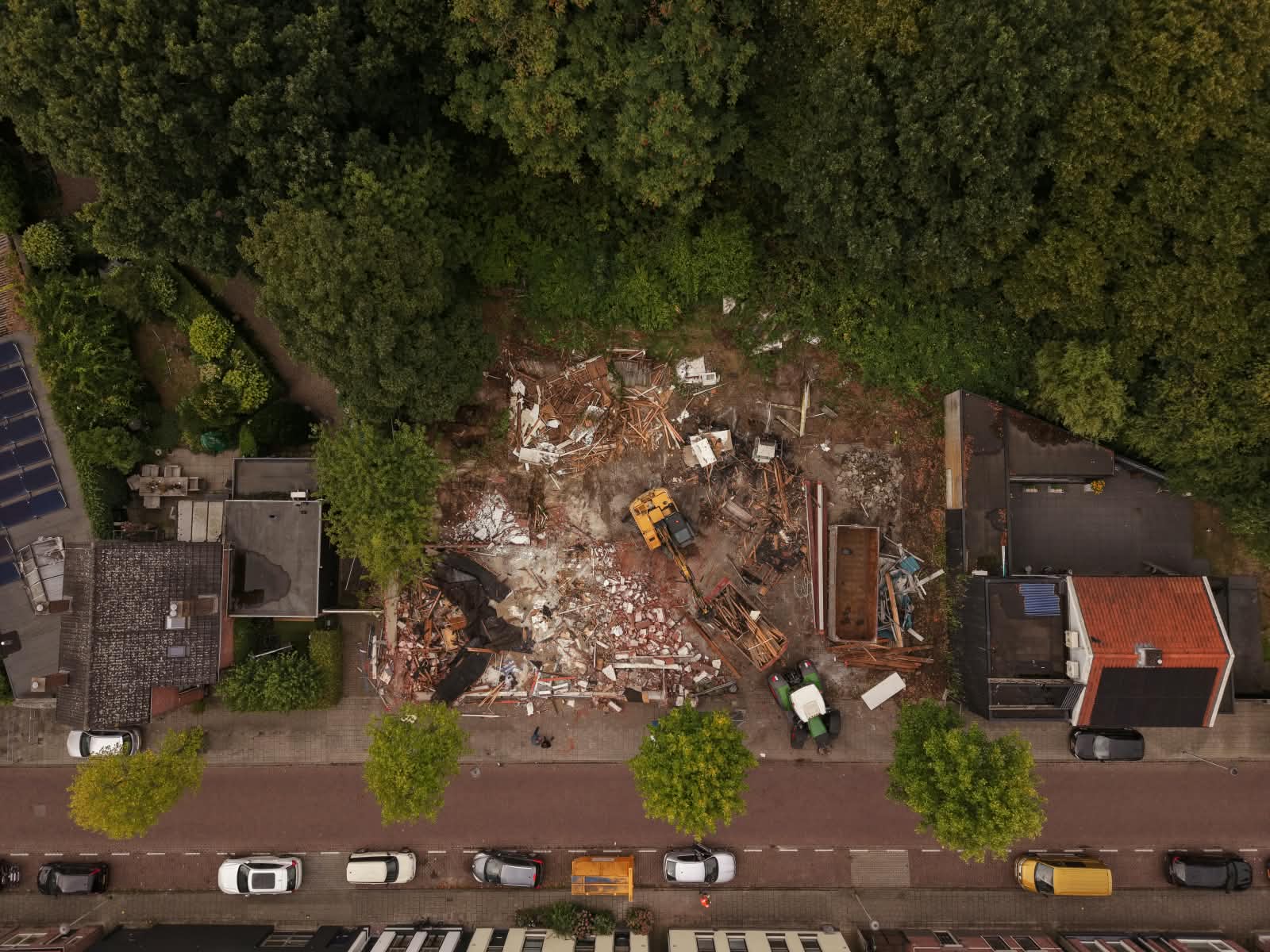 Luchtfoto van een bouwterrein met puin, houtafval, een graafmachine en een tractor, omgeven door bomen en huizen aan een straat met geparkeerde auto's.