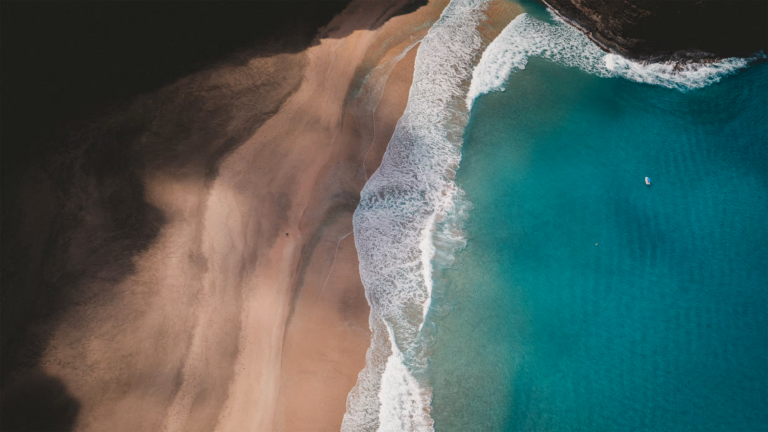 Aerial view of waves gently washing onto a sandy beach with turquoise ocean water and rocky shoreline.