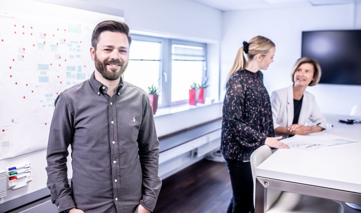 ein Mann steht im Raum, im Hintergrund ist eine Frau zu sehen, die arbeitet