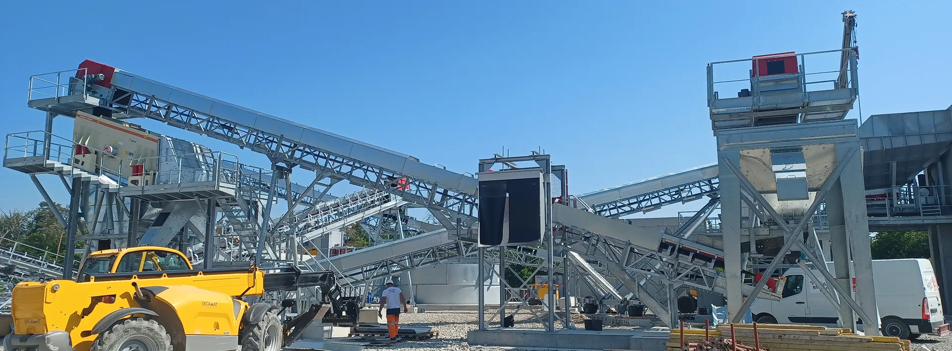 Site industriel de construction avec des structures de convoyage métalliques, une chargeuse jaune et un ouvrier portant un pantalon orange.