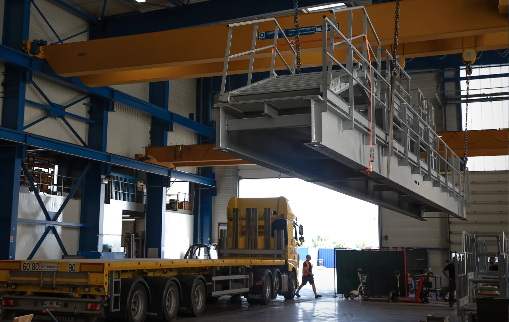 Grande plateforme métallique en cours de levage à l’intérieur d’un entrepôt, près d’un camion jaune et de travailleurs.