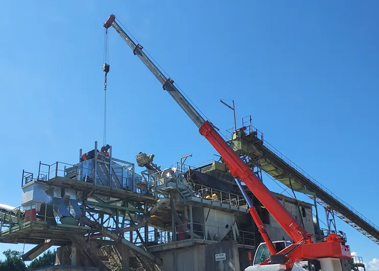 Grue rouge soulevant des matériaux sur un site industriel avec des structures métalliques et des convoyeurs sous un ciel bleu dégagé.