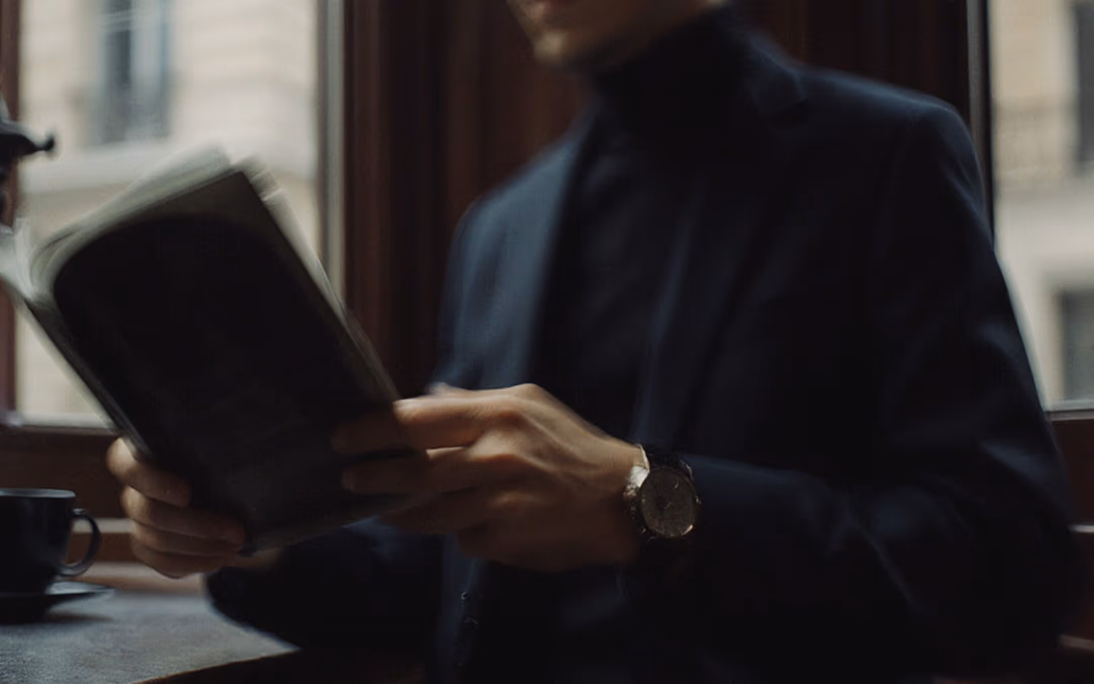 A sophisticated man in a dark suit and turtleneck reading a book by a window, wearing a high-value timepiece protected by Saef’s real-time security platform.