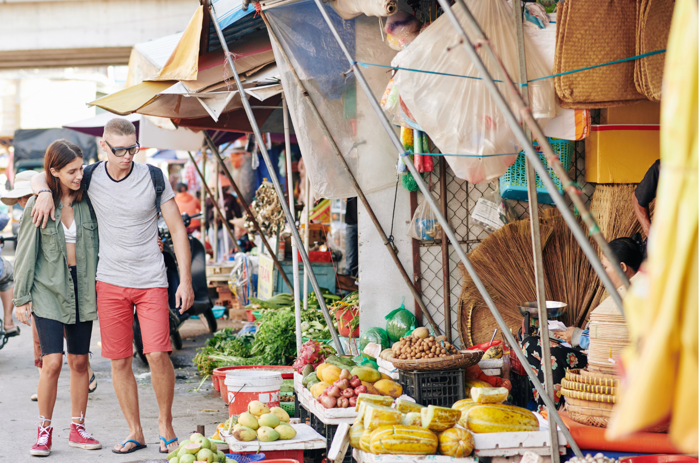 A young couple strolling leisurely through an Athenian street market, passing fruits, vegetables, and woven goods