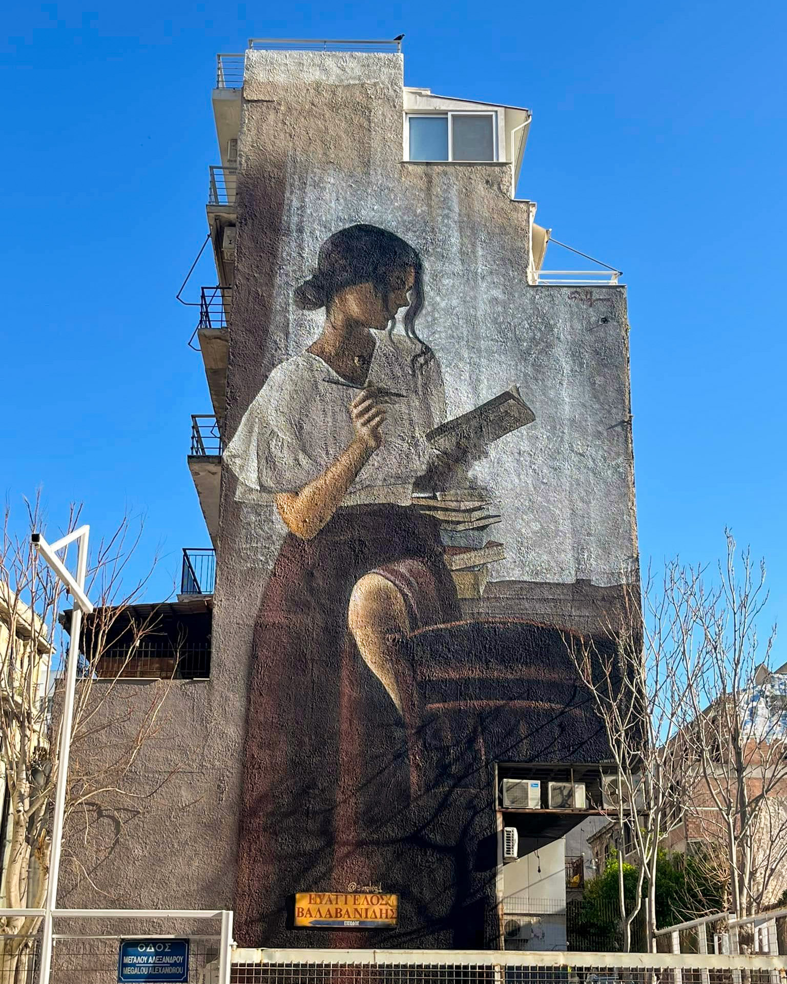 SimpleG mural of a woman reading beside stacked books on Megalou Alexandrou Street in Metaxourgeio, Athens.