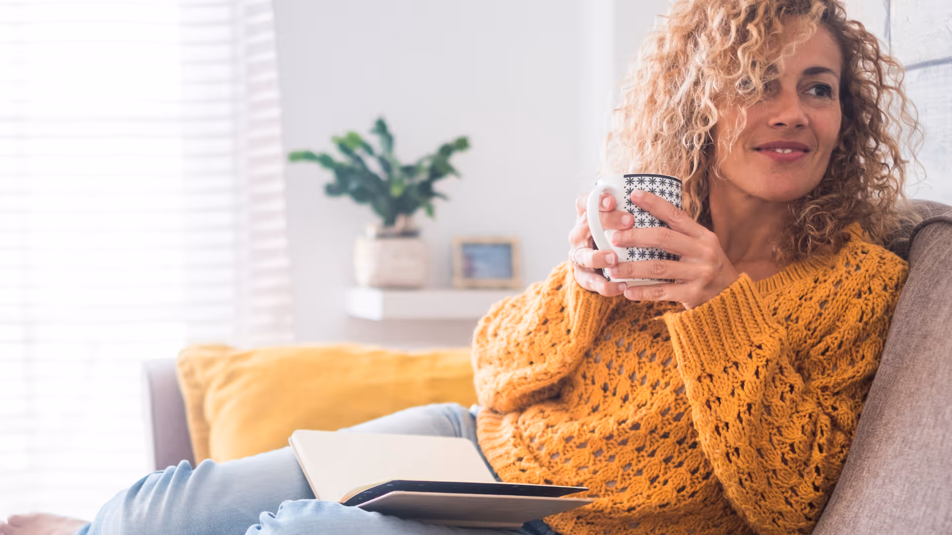 Smiling woman in a mustard sweater sitting on a couch with a mug and an open notebook on her lap.