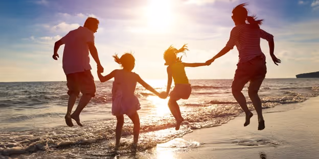Silhouettes of two adults and two children holding hands and jumping on the beach at sunset.