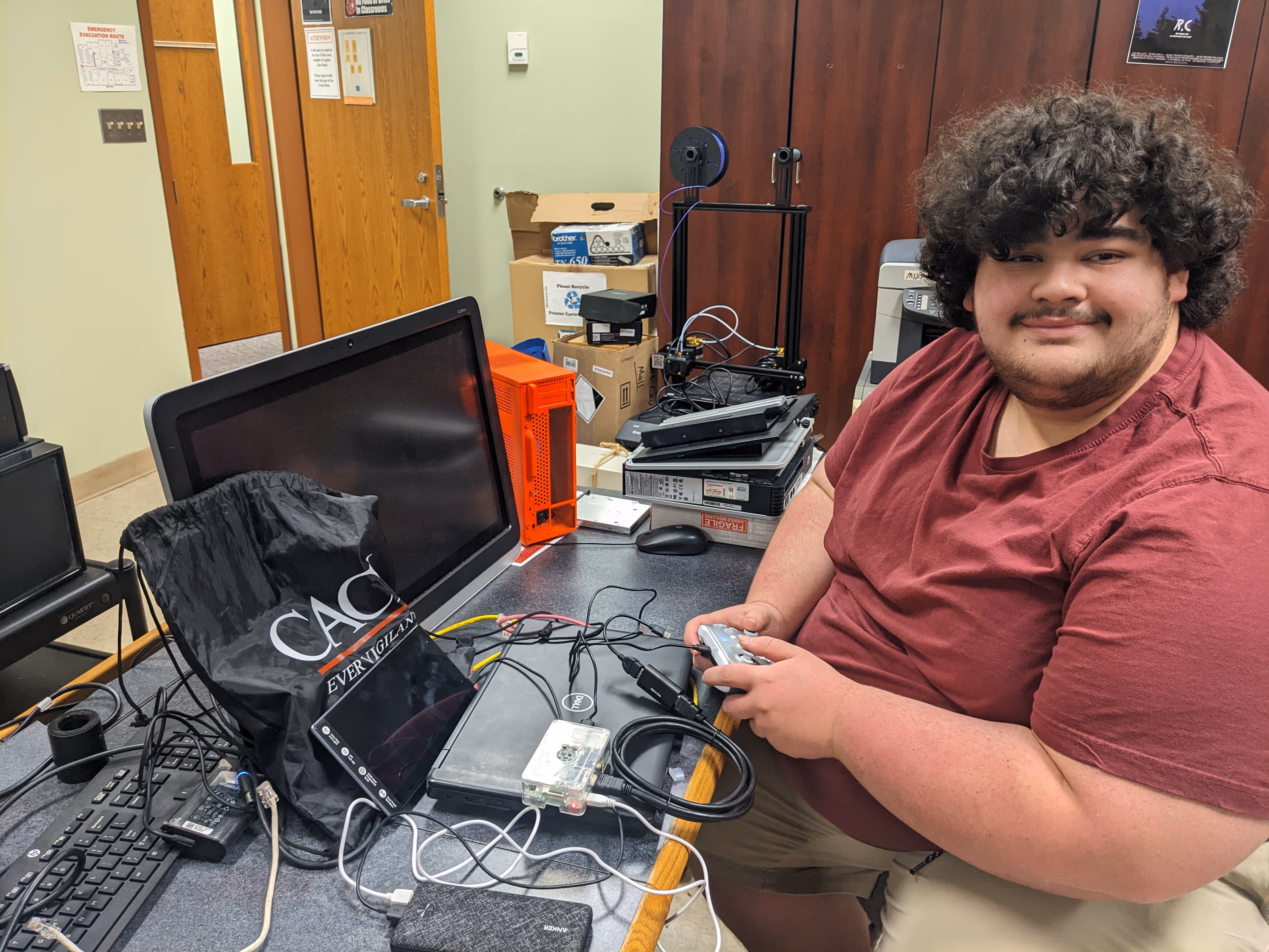 Man with curly hair in a maroon shirt sitting at a desk with monitors, computer parts, and a game controller in his hands.