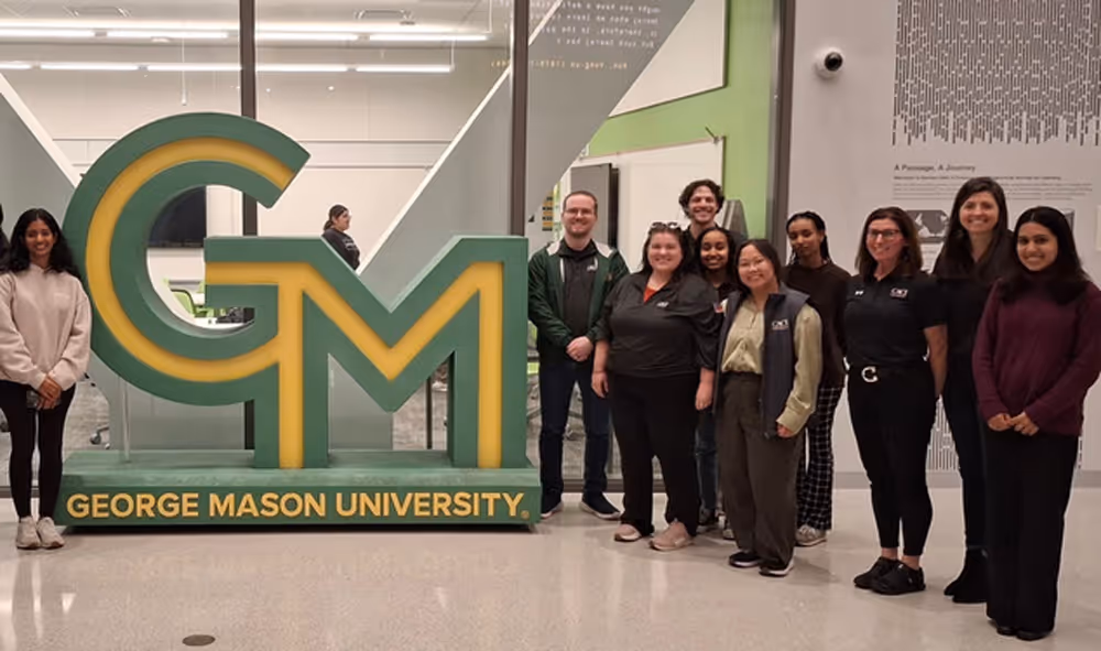Group of people standing indoors next to a large green and yellow sign that reads George Mason University.