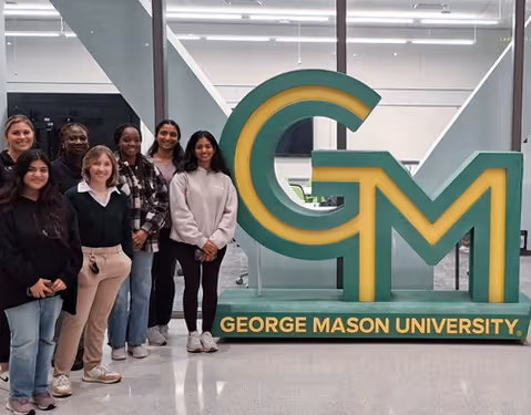Group of people standing indoors next to a large green and yellow sign that reads George Mason University.