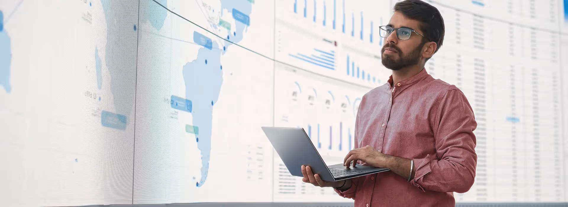 Man in glasses and red shirt holding a laptop and analyzing large data charts and maps on a screen.