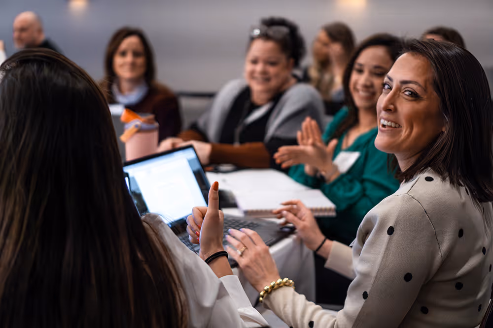 A group of diverse women sitting around a table, smiling and engaging in a discussion, with one woman giving a thumbs-up.