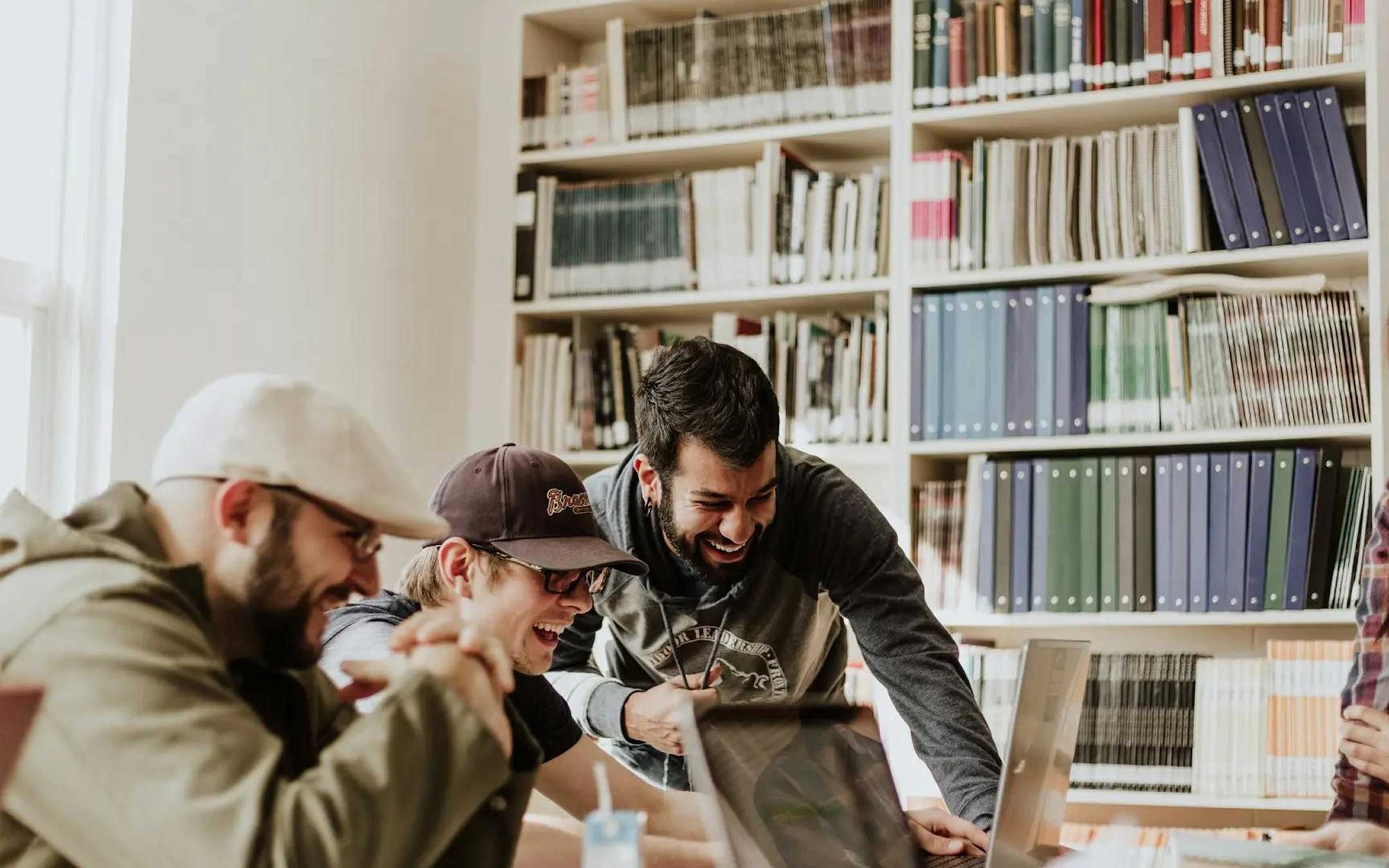 Trois jeunes hommes souriants et engagés devant des ordinateurs portables dans une bibliothèque avec des étagères remplies de livres et dossiers.