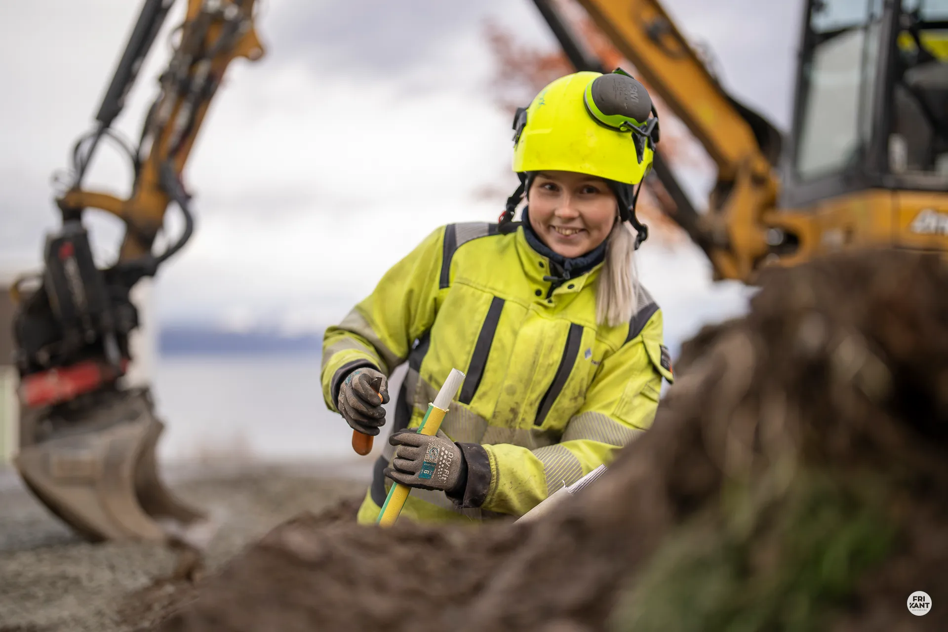Kvinne i gul hjelm og refleksjakke arbeider med kabel på en byggeplass med gravemaskin i bakgrunnen.