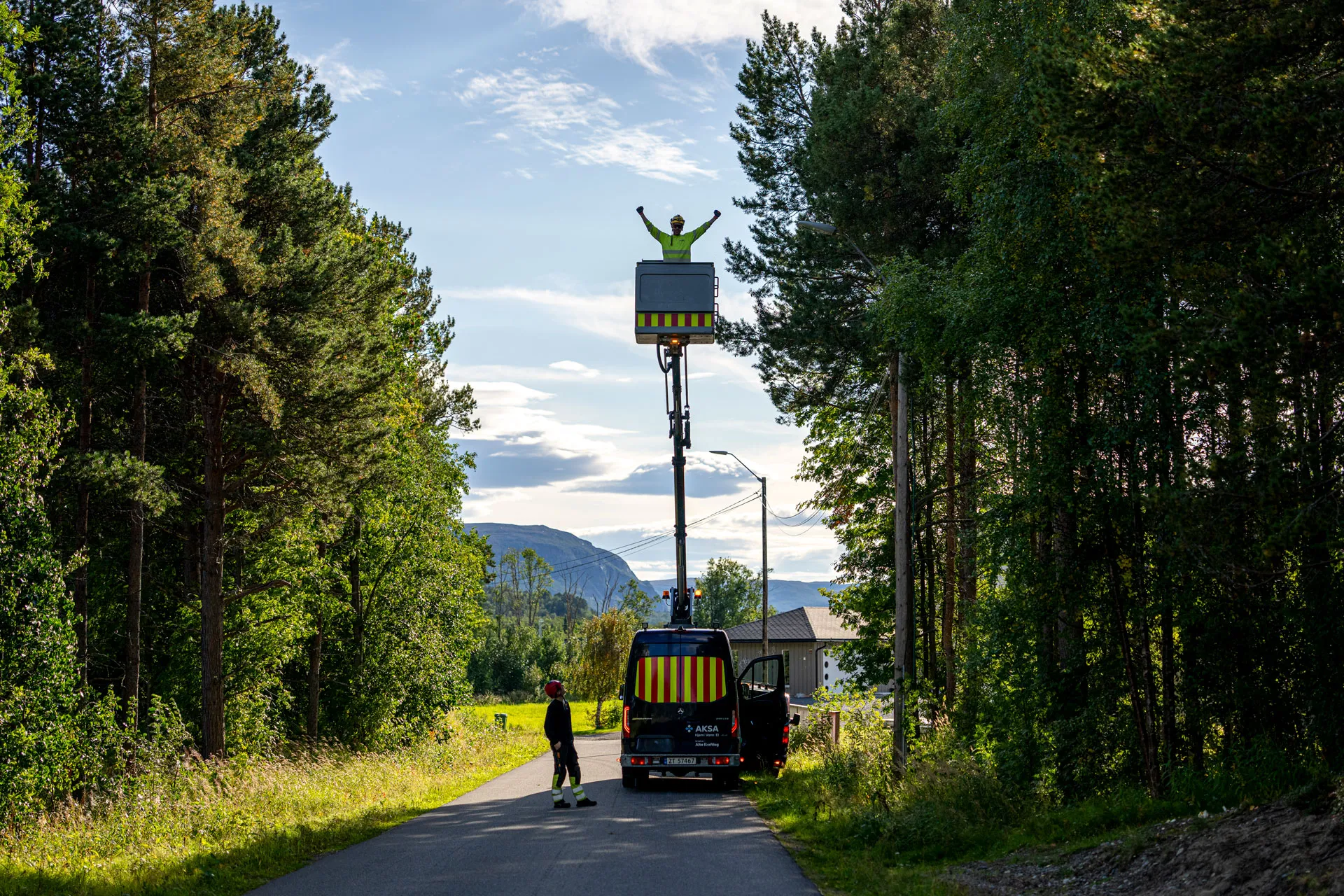 To arbeidere, en i en heisarm løftet fra en varebil på en landevei omgitt av trær under en blå himmel.