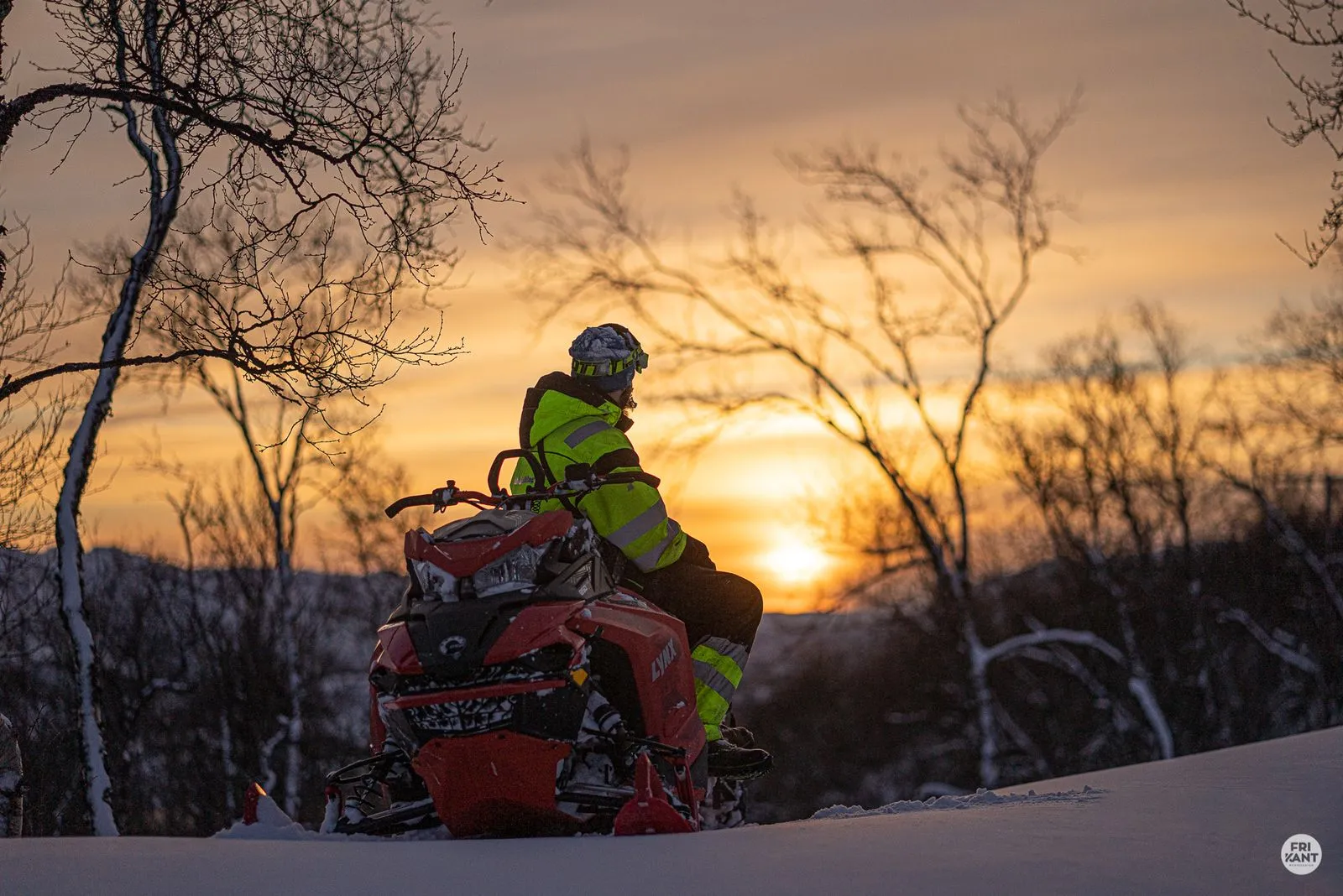 En person kledd i refleksdrakt sitter på en snøskuter og ser mot solnedgangen i et vinterlandskap med nakne trær.