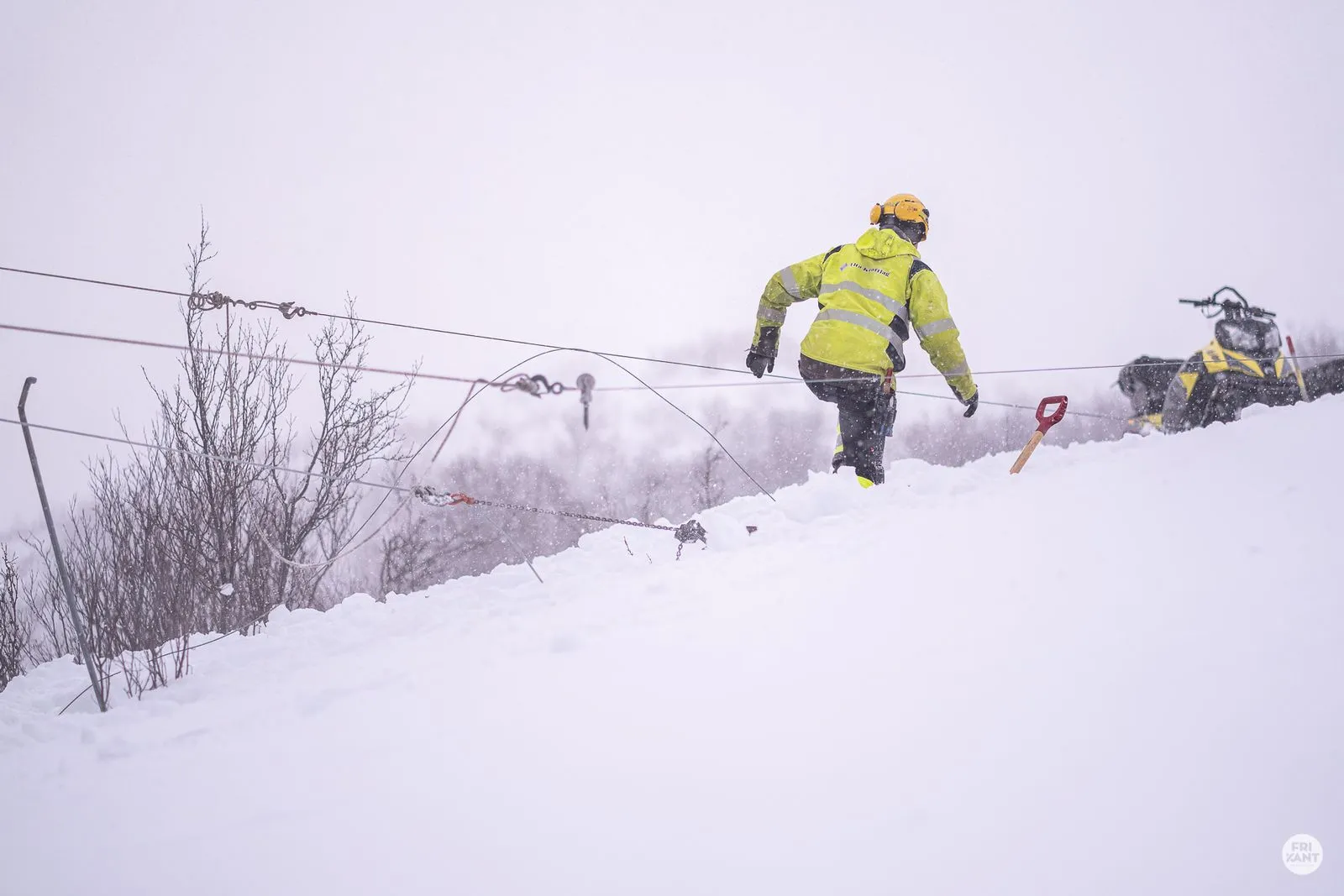 En person i gul refleksjakke og hjelm jobber i snøen ved en snøscooter og en rød snøskuffe under snøfall.