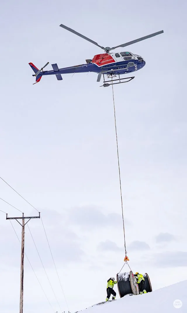Helikopter løfter en stor kabeltrommel mens to arbeidere i verneutstyr styrer den på en snødekt bakke.