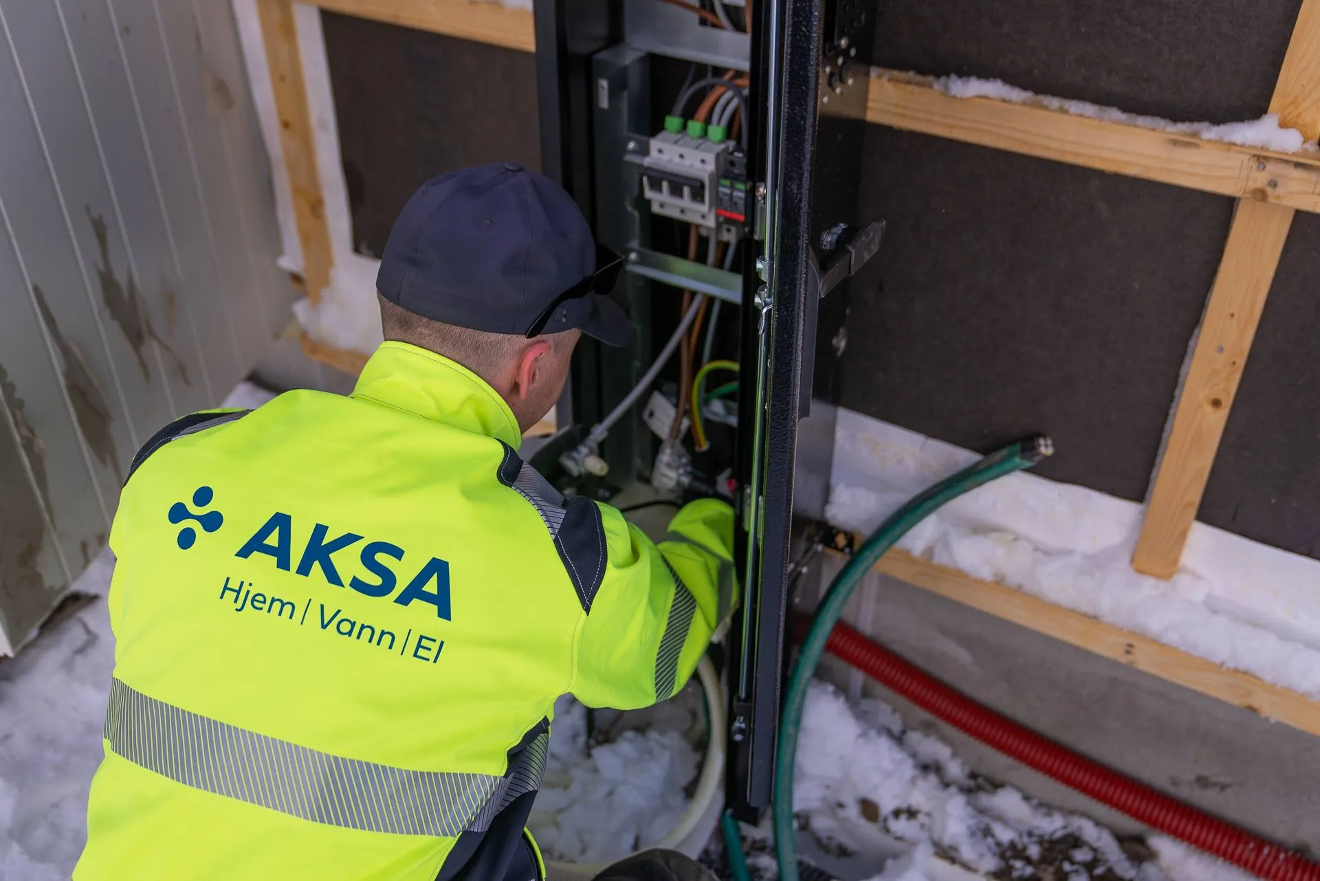 Tekniker i gul jakke med AKSA-logo jobber med elektrisk utstyr utendørs om vinteren.