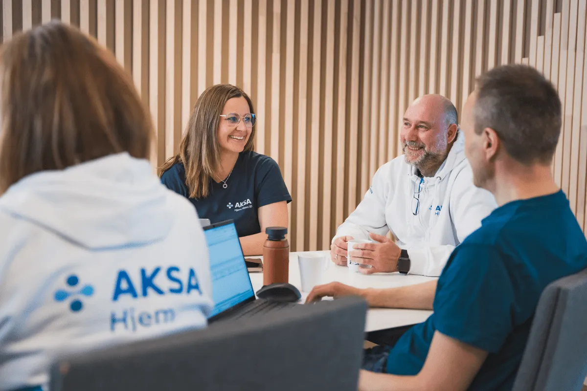 Fire personer i AKSA Hjem-klær sitter rundt et bord og har en hyggelig samtale i et moderne rom.