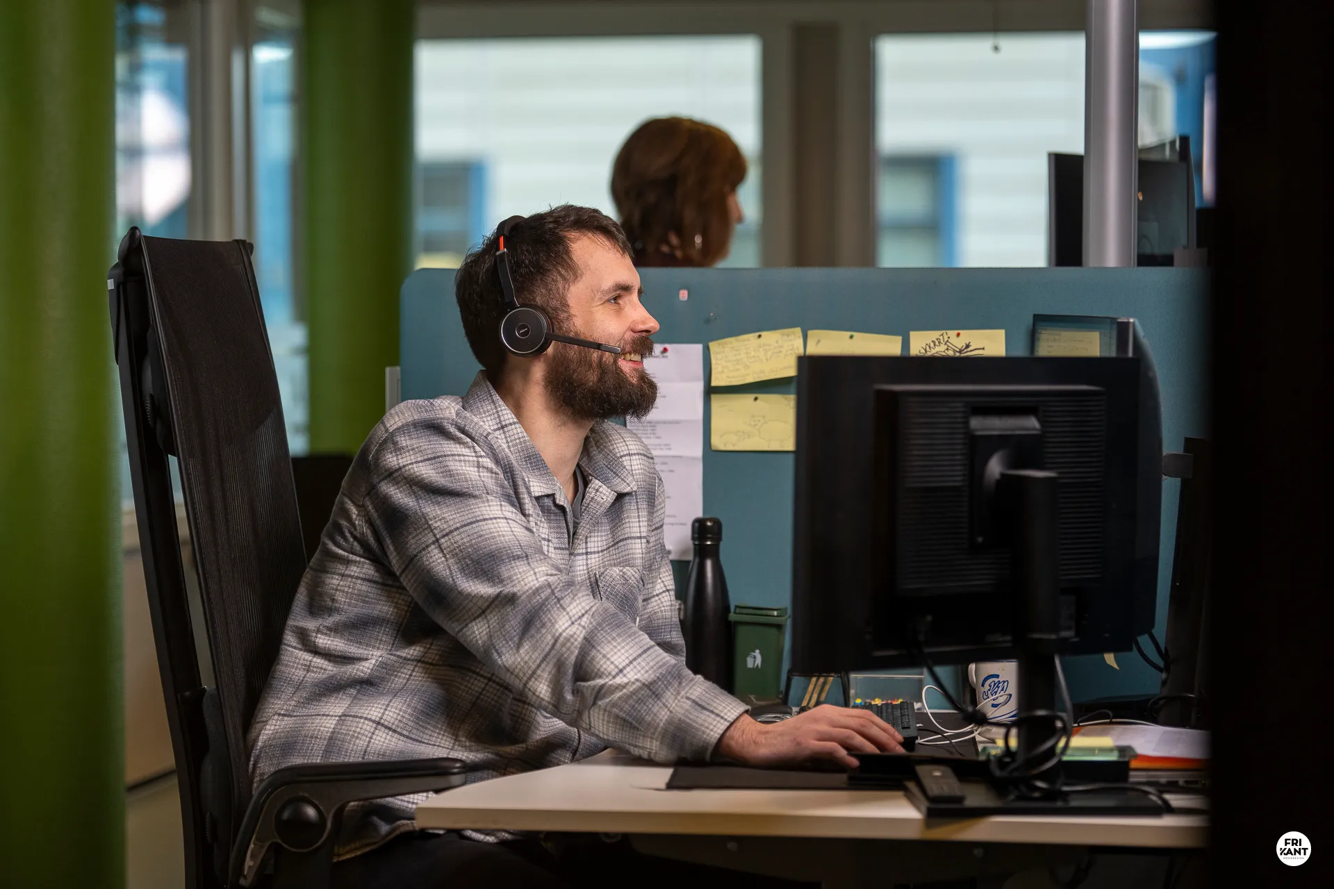 Smilende mann med headset som jobber ved datamaskin i kontorlandskap.