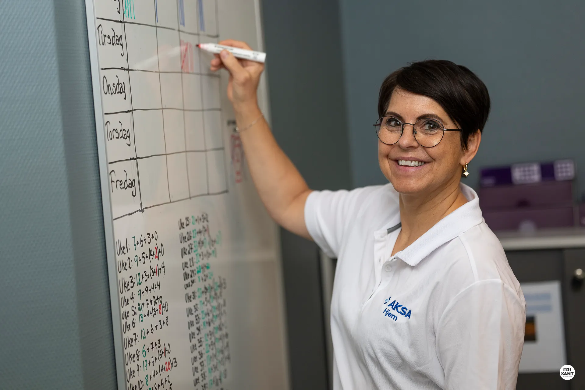 En kvinne med kort hår og briller smiler mens hun skriver med en rød tusj på en tavle med en ukesplan.