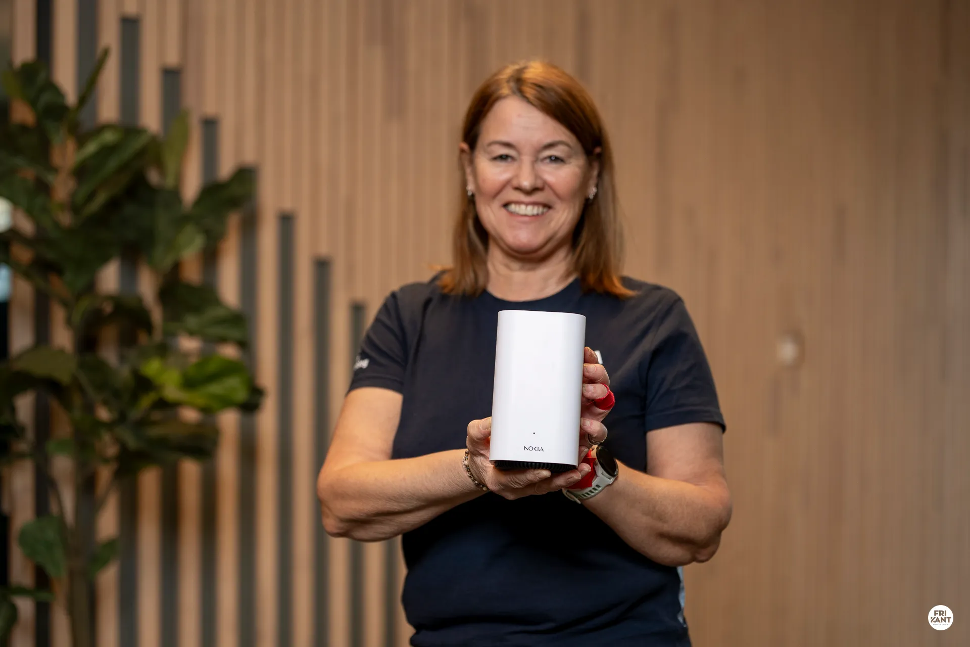 Kvinne smilende holder en hvit Nokia-enhet foran seg med begge hender, med en bakgrunn av trepaneler og en grønn plante.