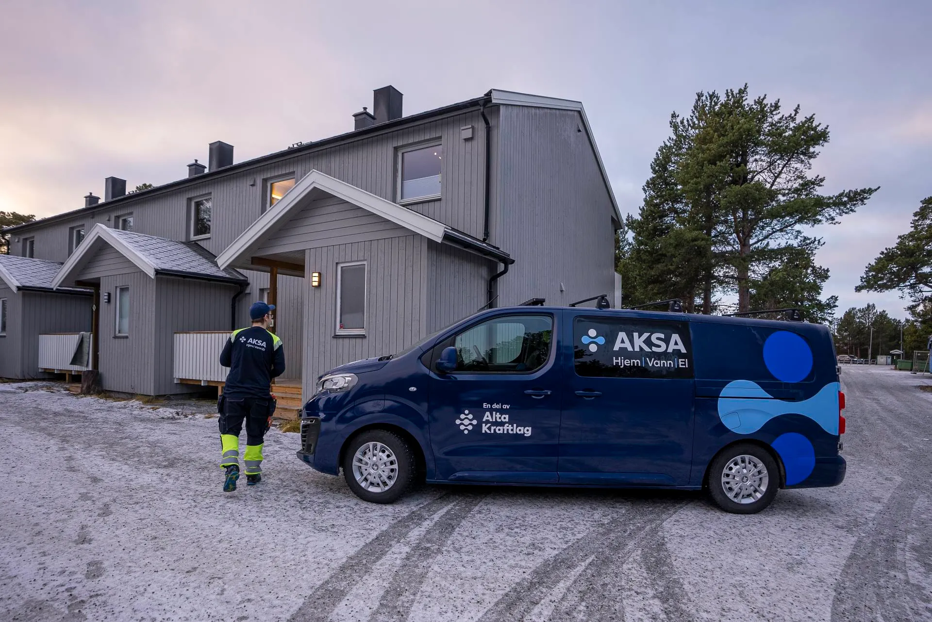 Tekniker går mot inngangen til en grå bolig med en mørk blå varebil merket AKSA parkert utenfor på en snødekt vei.