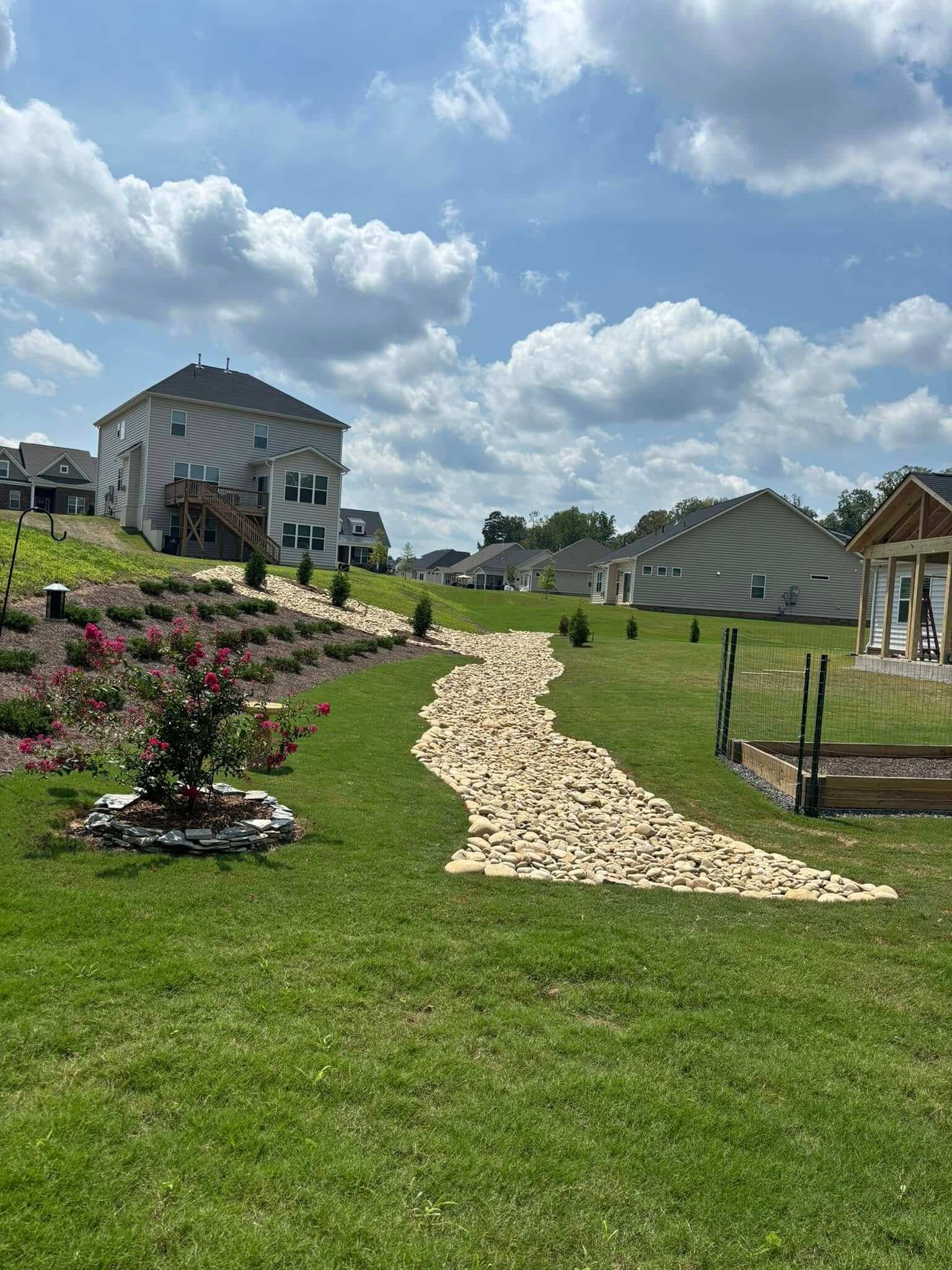 Residential neighborhood with a large green lawn, a dry creek bed with light-colored stones, flowering shrubs, and houses under a partly cloudy blue sky.