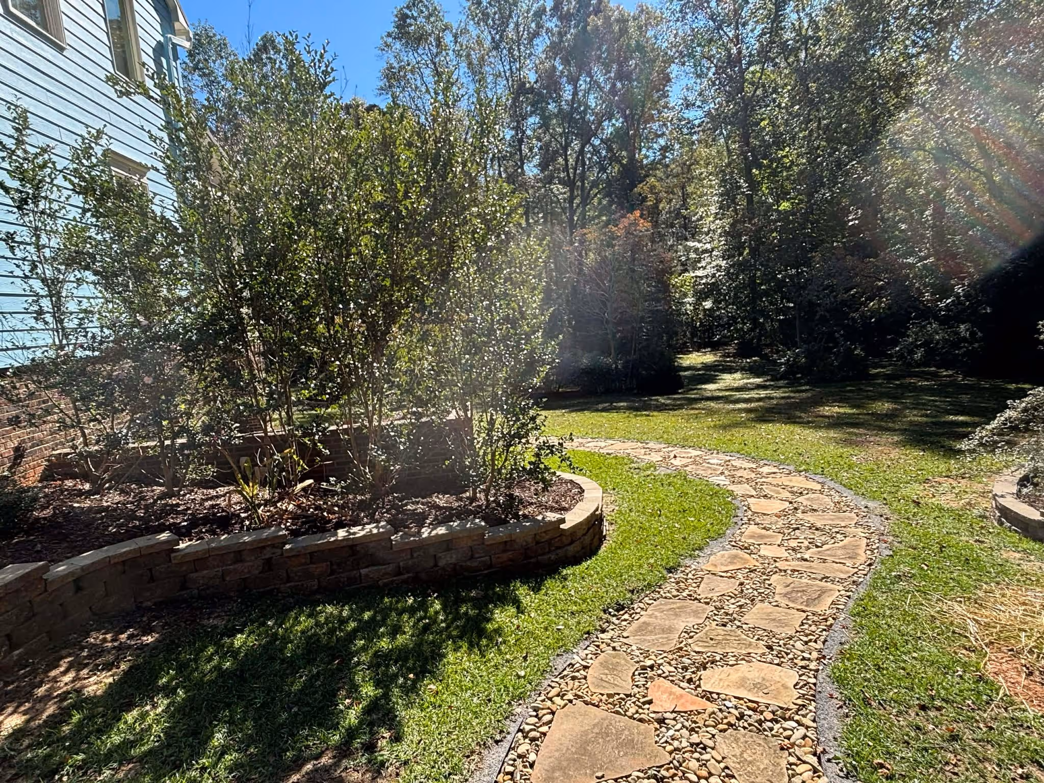 Curved stone and pebble pathway in a sunny backyard with green grass and shrubs beside a white house.