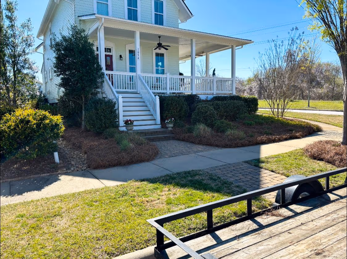 White two-story house with front porch, stairs, and surrounding bushes under a clear blue sky.