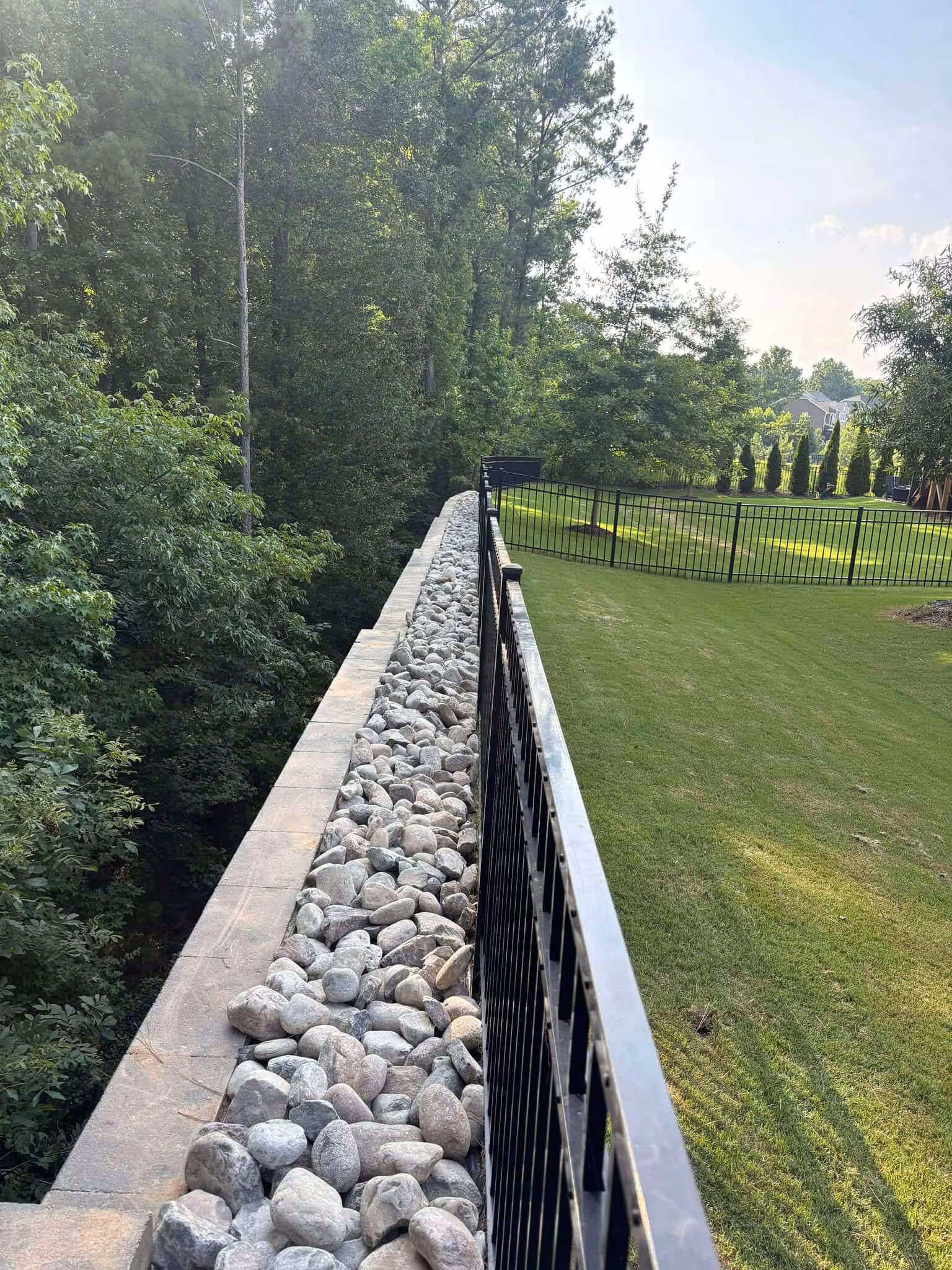 Stone-lined retaining wall with metal railing separating a grassy yard from a wooded area.