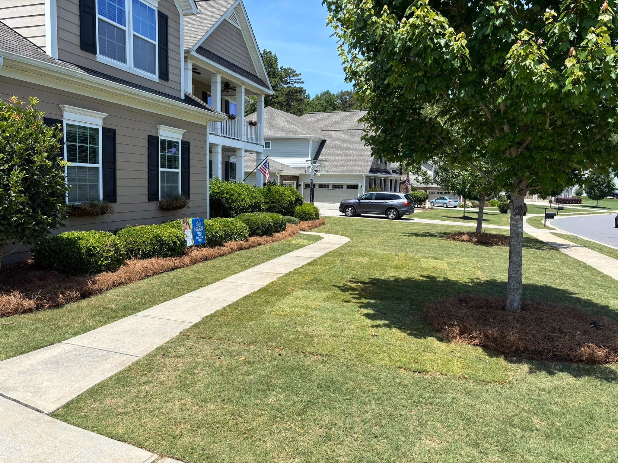 Suburban street view with tan house with black shutters, trimmed bushes, a lush green lawn, and a tree casting a shadow on the grass.