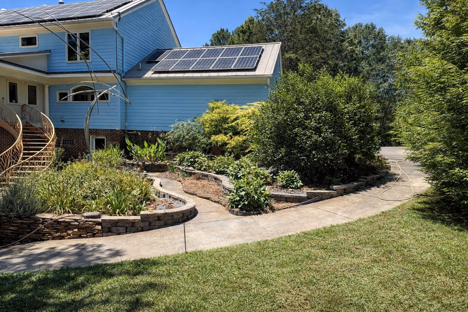 Blue house with solar panels on roof, stone-bordered garden beds, and a curved concrete pathway surrounded by green bushes and trees.