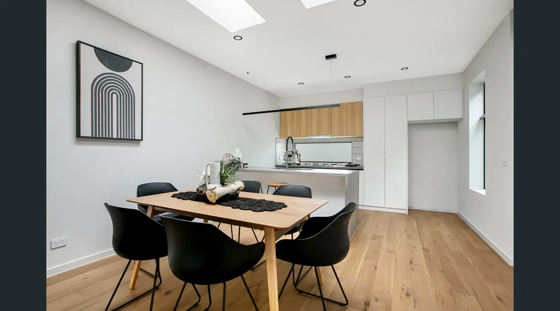 Modern dining area with wooden table, six black chairs, minimalist kitchen with white cabinets and wood accents, and skylights overhead.