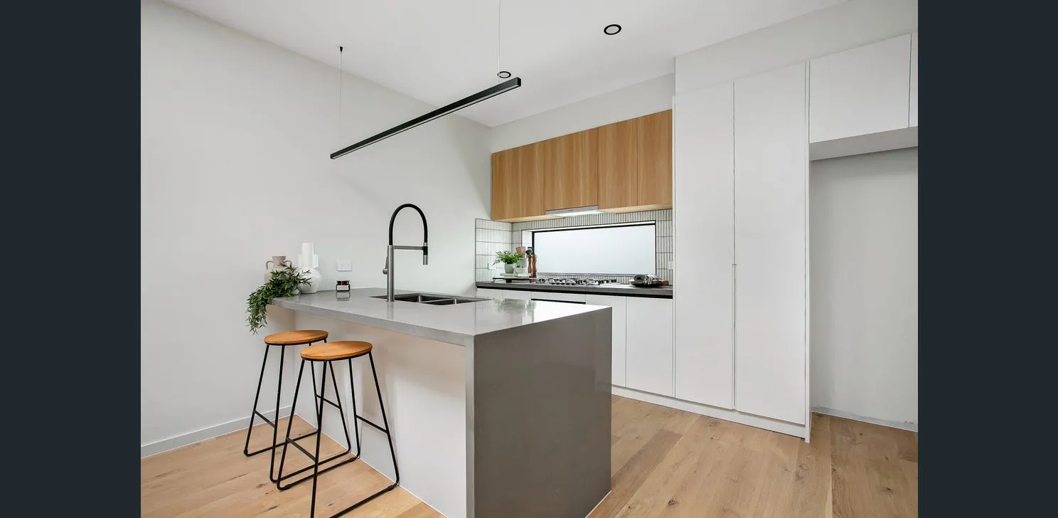 Modern kitchen with a gray island counter, two wooden stools, white cabinets, and wooden upper cupboards with a large window backsplash.