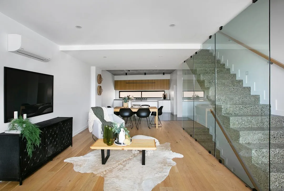 Modern open living space with wooden floor, gray armchair, wooden coffee table on fur rug, dining table with black chairs, and terrazzo staircase with glass railing.
