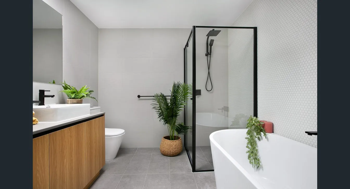 Modern bathroom with a wooden vanity, white sink, toilet, glass shower with black frame, freestanding bathtub, and decorative green plants.