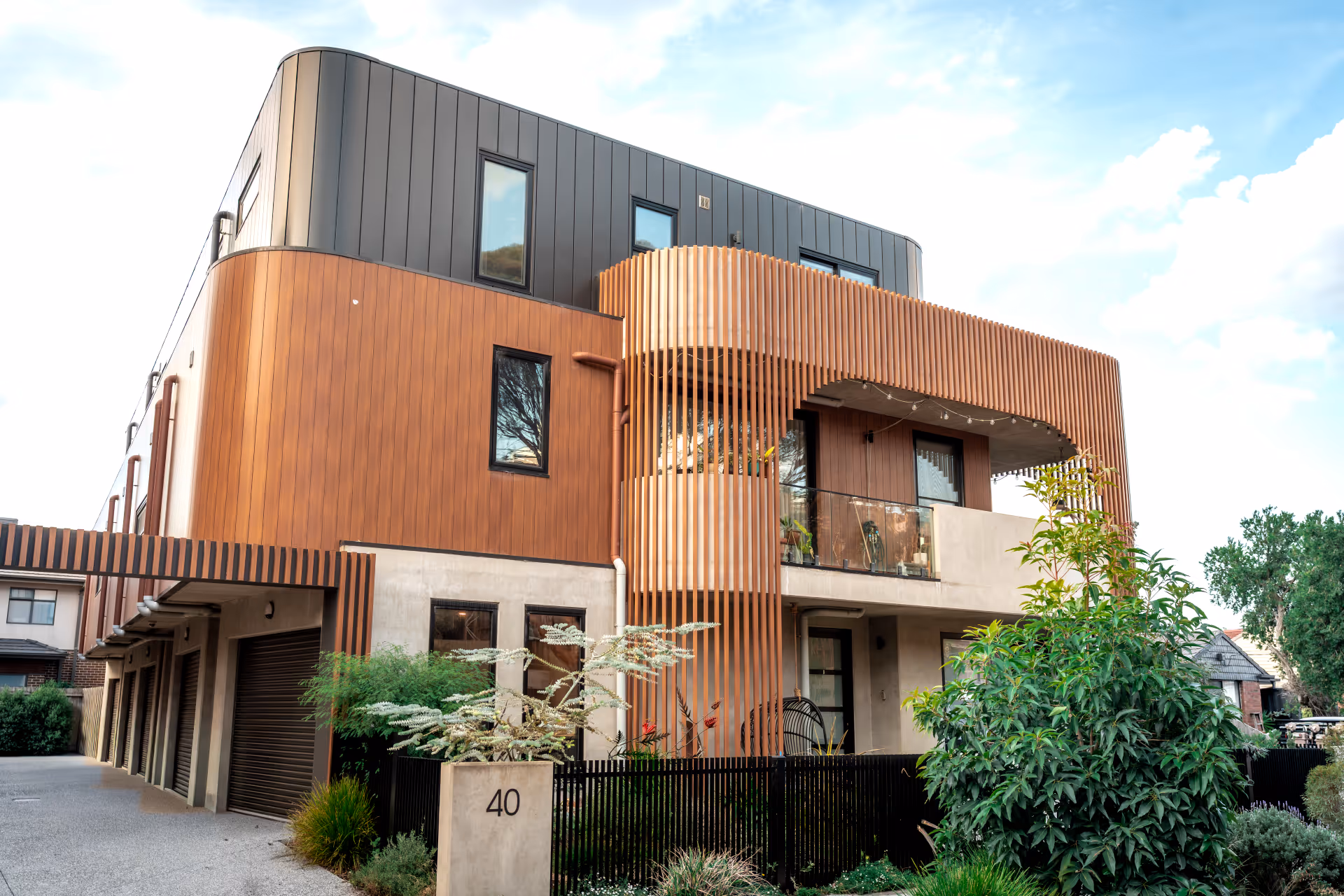 Modern multi-story residential building with wood paneling, rounded balcony with vertical slats, and landscaped front garden.