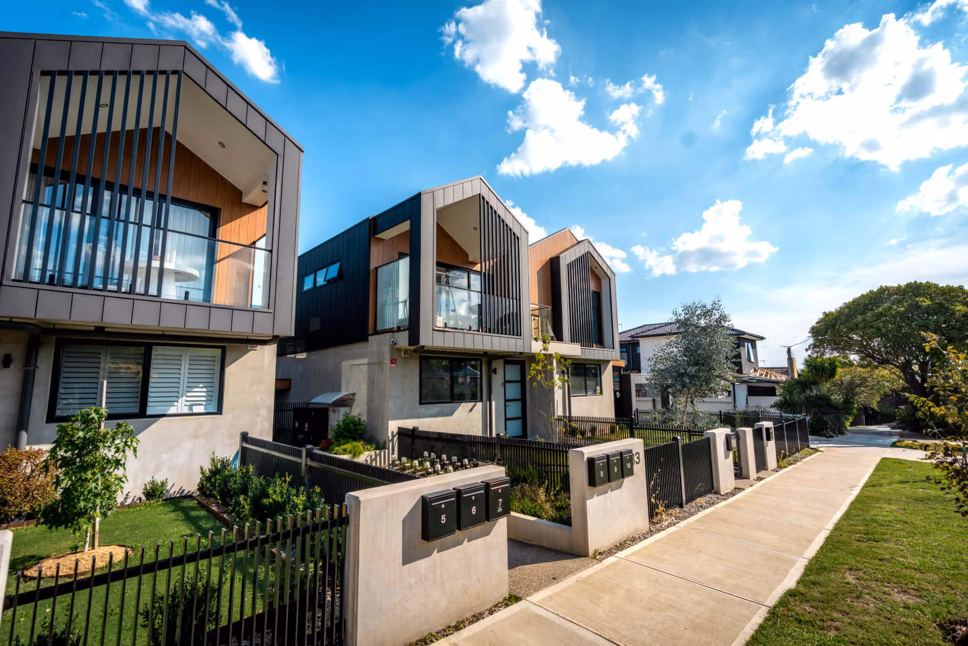 Modern townhouses with large windows, balconies, and fenced front yards along a sidewalk under a partly cloudy sky.