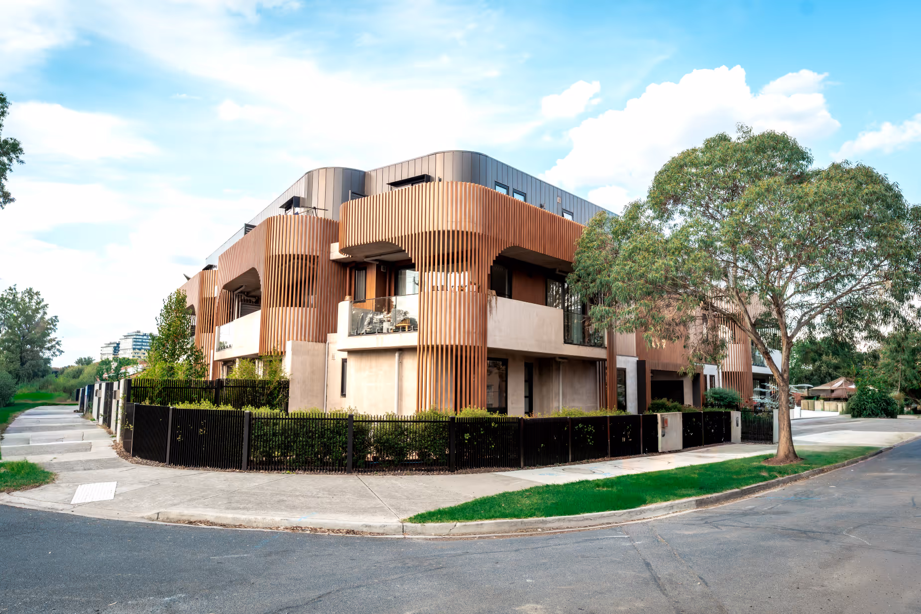 Modern corner building with vertical wooden slats, large balconies, and surrounding black fence under a blue sky.
