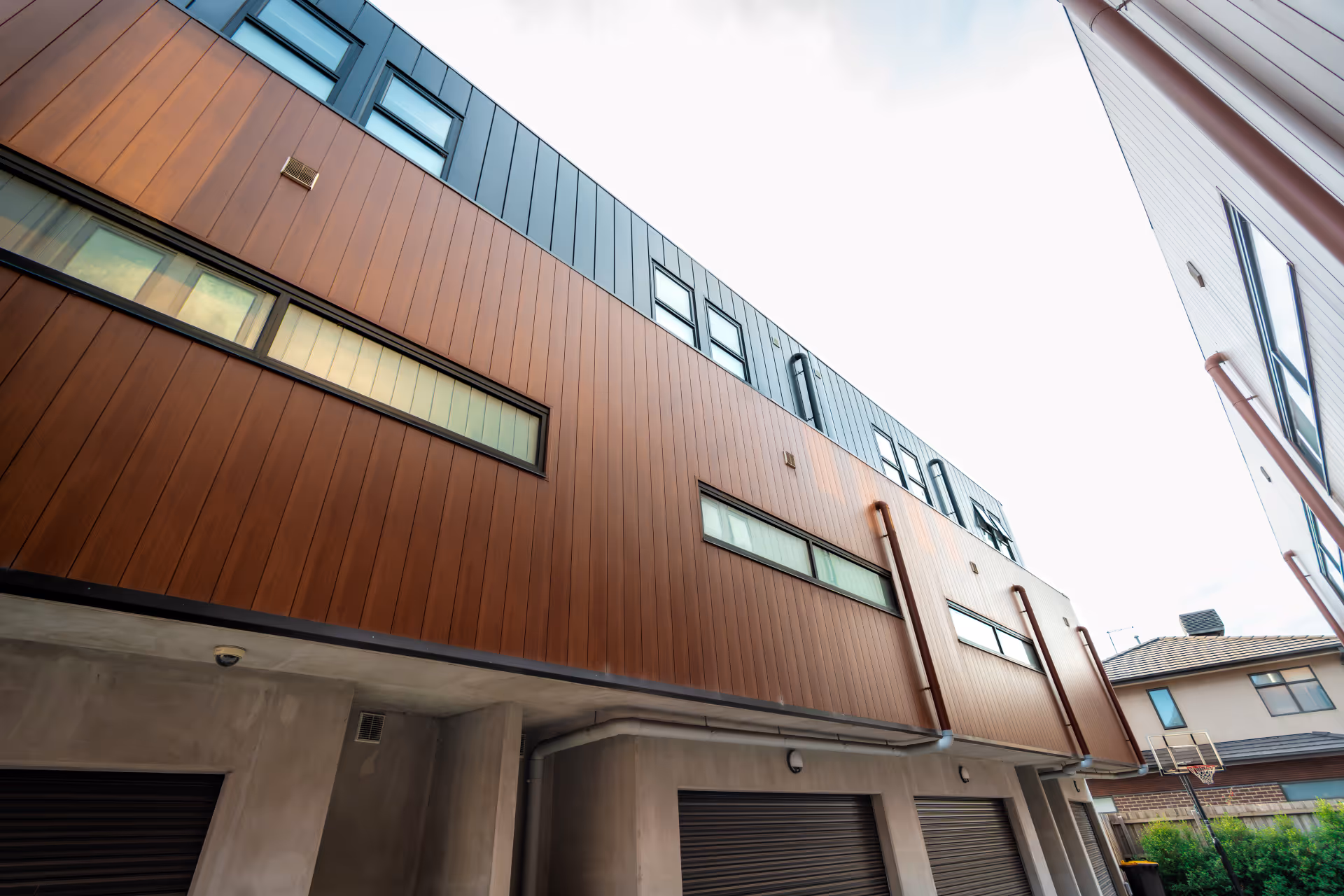 Modern multi-story residential building with brown and blue cladding, narrow horizontal windows, and garage doors on the ground level.