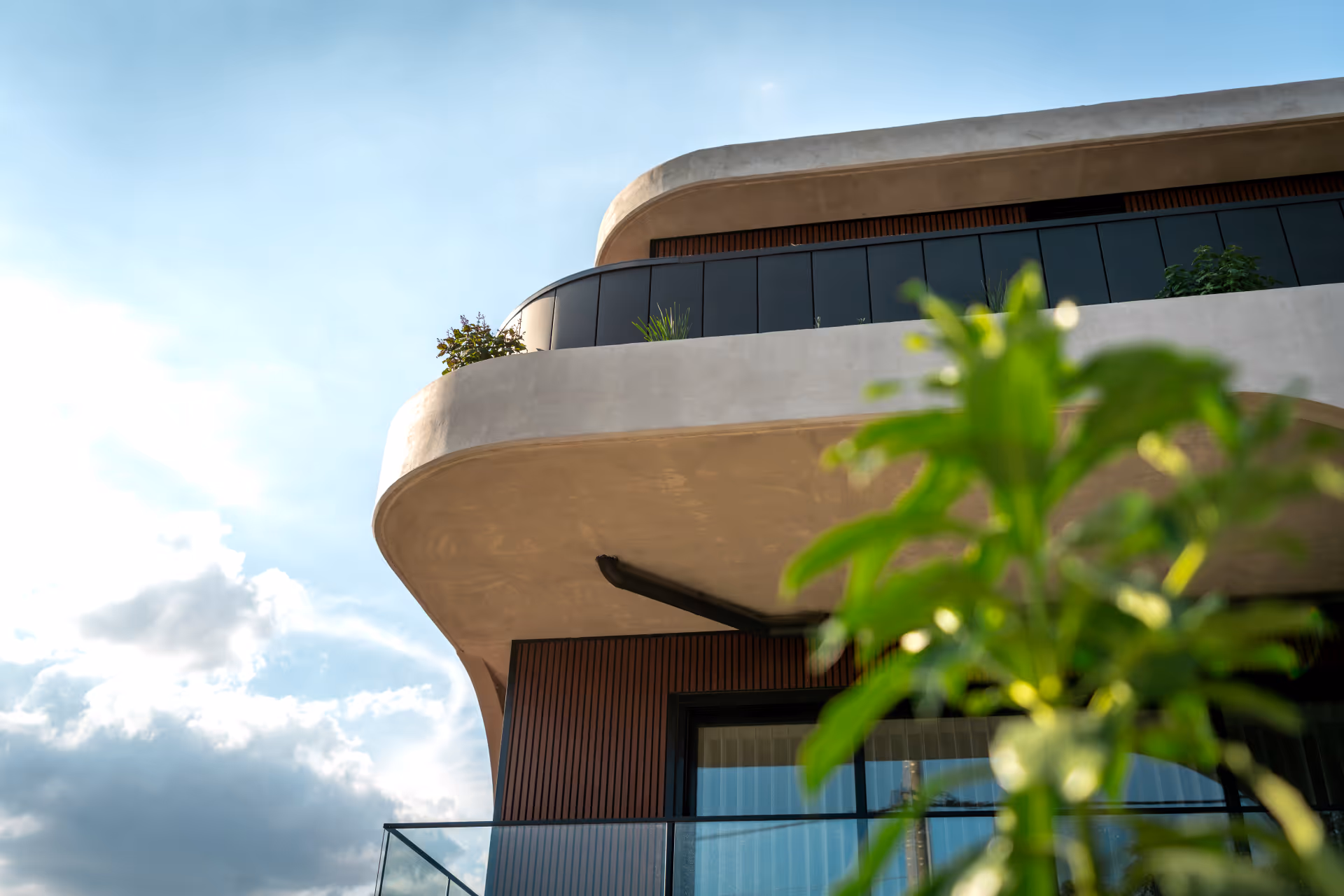 Modern curved balcony with glass railing and potted plants under blue sky and clouds.