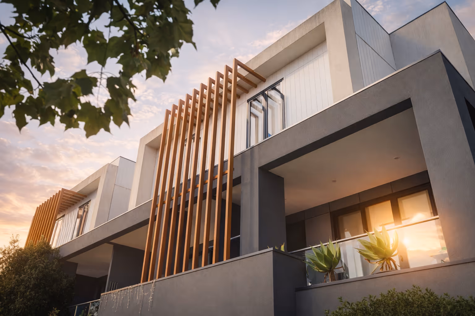 Modern two-story building with vertical wooden slats, large windows, and plants on the balcony at sunset.