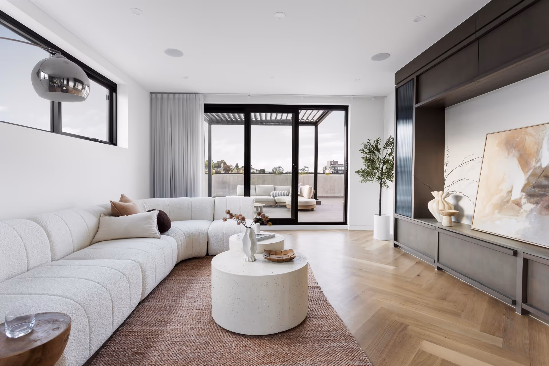 Modern living room with a curved white sofa, round coffee table, large sliding glass doors leading to an outdoor patio, and a built-in dark wood entertainment unit.