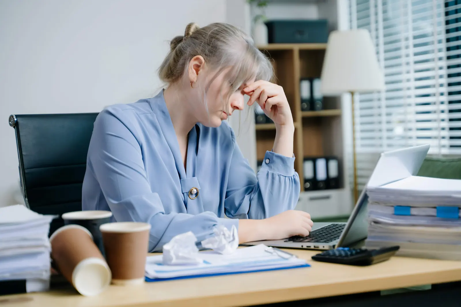 Exhausted woman holding her head at a cluttered office desk with a laptop and coffee cups.