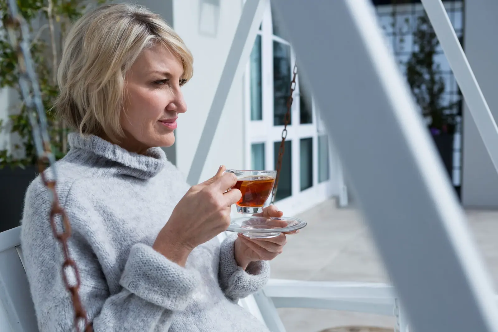 Woman in a grey jumper drinking a cup of tea on a porch swing.
