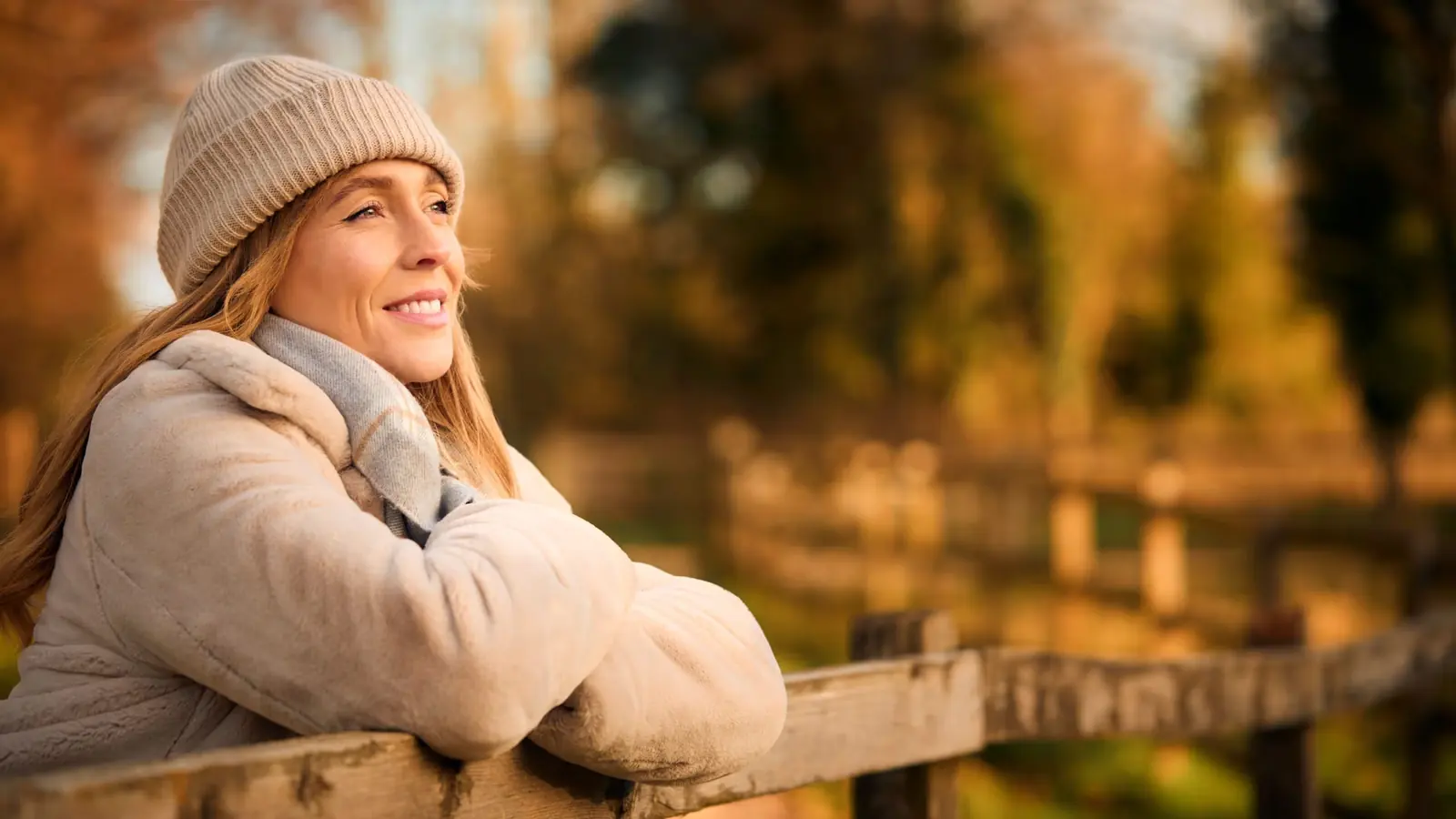 Smiling woman in a woolly hat leaning on a fence in an autumn park.