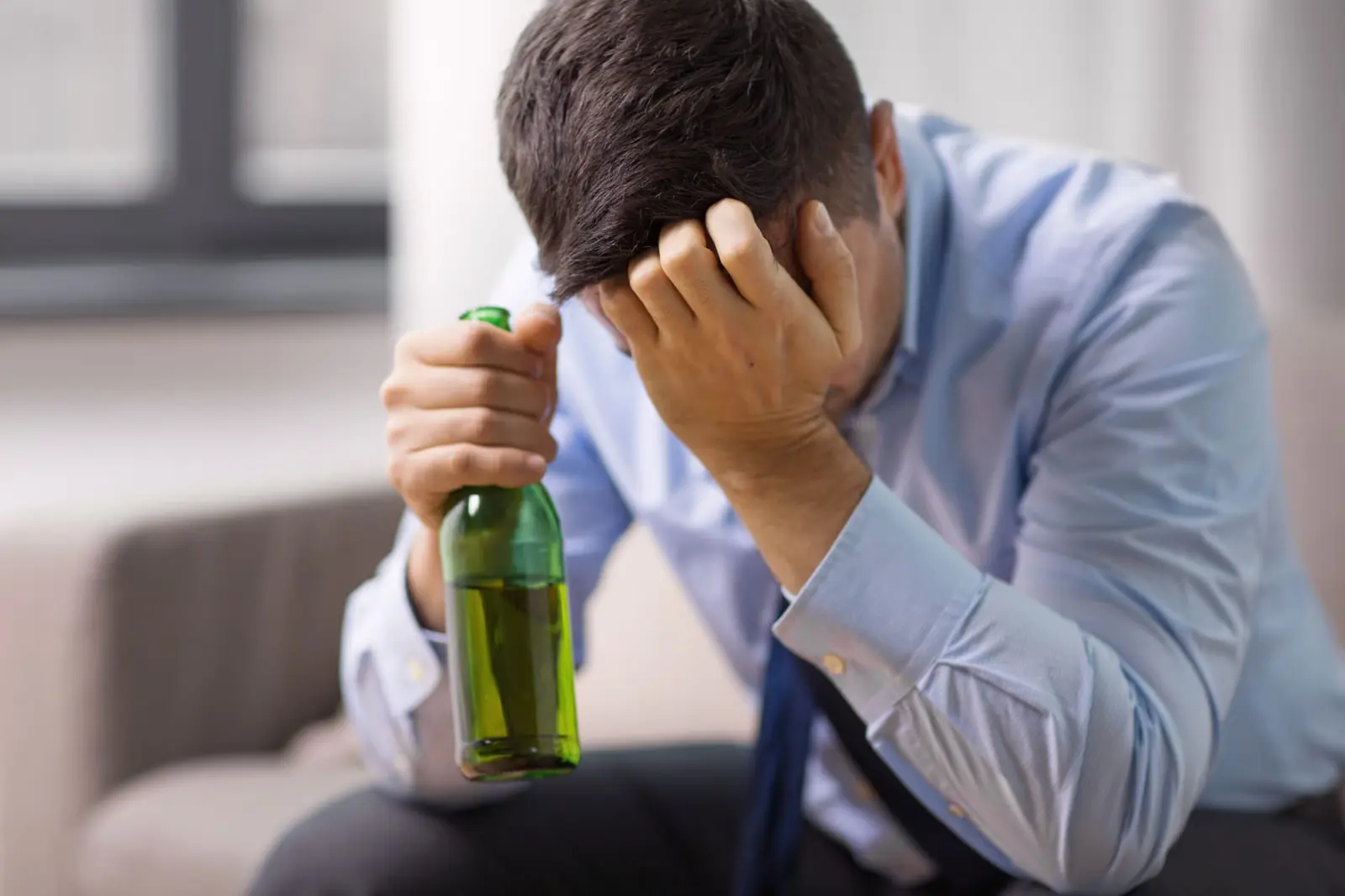 Man in a shirt and tie holding his head in one hand and a bottle of beer in the other.